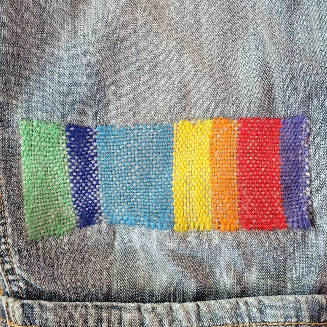 A woven patch on blue denim. The darn was woven in a loose rainbow pattern, switching between vertical blocks of colour from green, through several shades of blue, to yellow, orange, red, and purple.